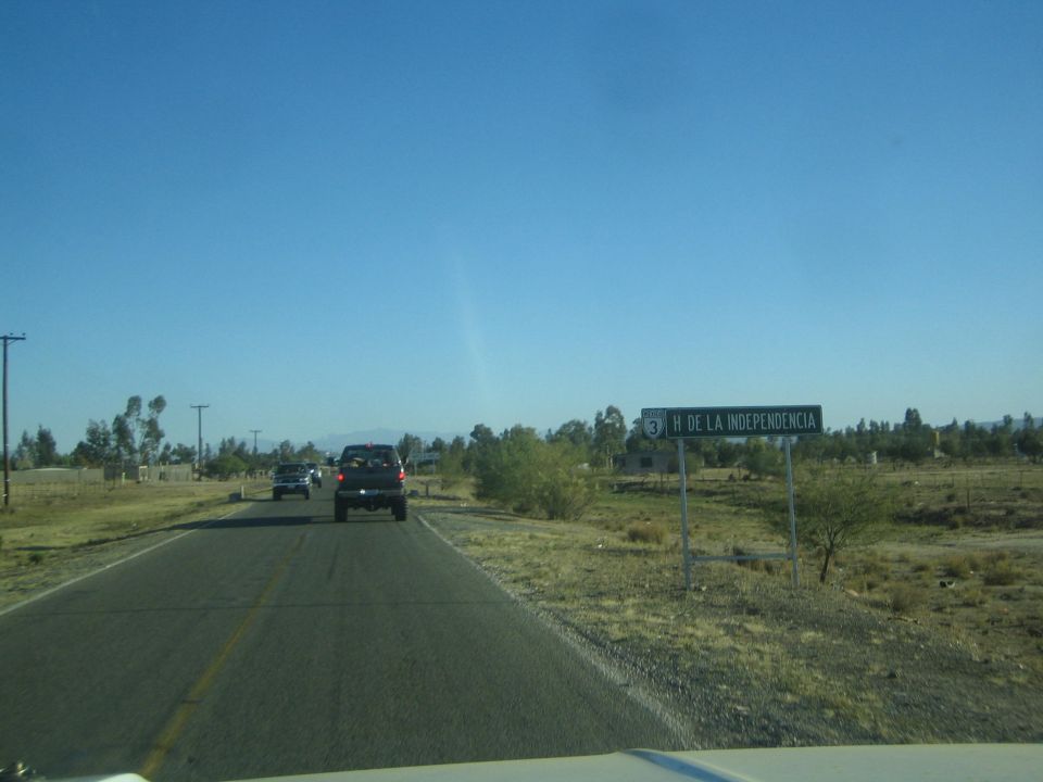 Rural road with a roadside sign reading H DE LA INDEPENDENCIA and vehicles ahead