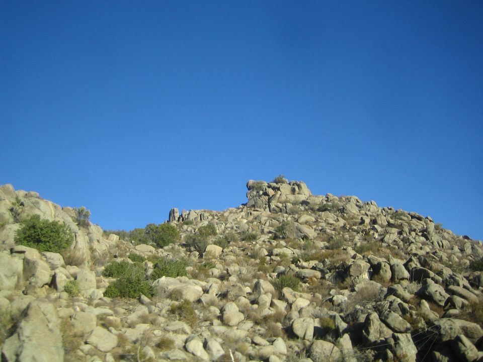 Rocky boulder-strewn hillside with sparse green shrubs under a clear blue sky