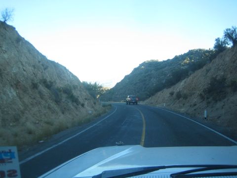 1990 Ford Bronco - Seen through a windshield, a paved road winds through a cut between brushy hillsides at dusk with a pickup truck driving ahead.