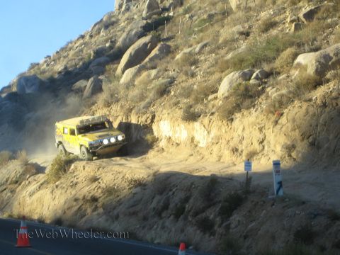 1990 Ford Bronco - yellow off-road race truck kicks up dust as it drives along a rugged dirt embankment beside a paved road in golden light.