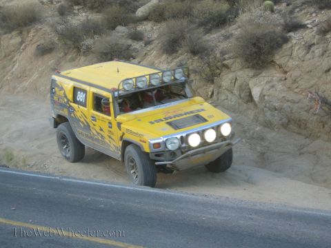 1990 Ford Bronco - yellow Hummer H2 race truck numbered 861, with headlights on and a roof light bar, climbs a rocky hillside trail above a road.