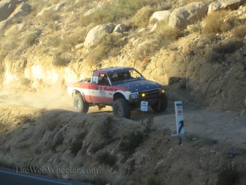 1990 Ford Bronco - red, white and blue single-cab prerunner race truck with yellow fog lights drives a dirt trail above a paved road at dusk.