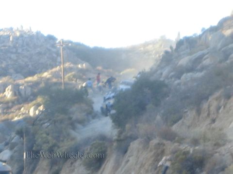 1990 Ford Bronco - hazy, dust-filled scene shows several people and an off-road vehicle on a steep dirt trail across a rocky hillside at dusk.