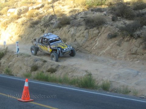 1990 Ford Bronco - black off-road race buggy with yellow flame graphics and a roof light bar drives a dirt trail above a paved road marked by a cone.