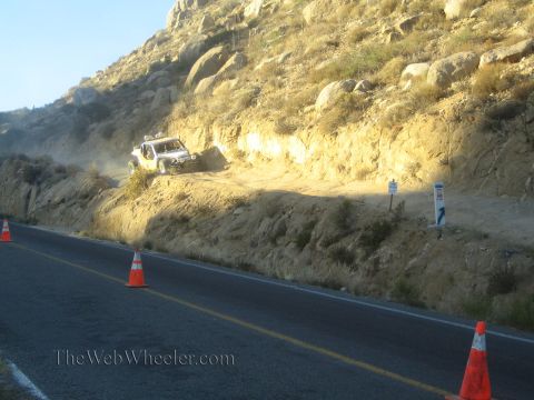 1990 Ford Bronco - white off-road race truck raises dust on a steep dirt embankment above a paved road lined with orange traffic cones at dusk.