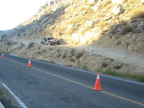 1990 Ford Bronco - dark off-road vehicle sits on a dirt trail above a paved road marked by a line of orange traffic cones beneath a rocky hillside.