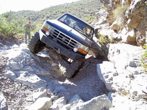 1991 Ford Bronco - A dark blue Ford Bronco navigates a challenging rocky trail, demonstrating its off-road capabilities.