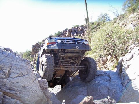 1991 Ford Bronco - Ford Bronco with aftermarket bumper and large tires crawling over rocks on an off-road trail.