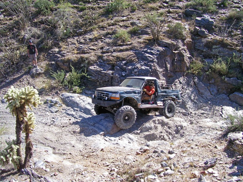 Pickup truck with large tires navigating a rocky, desert trail. Driver is visible