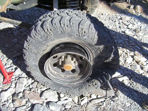 1991 Ford Bronco - A flat tire is shown on a vehicle, with a red jack positioned nearby on the rocky ground.