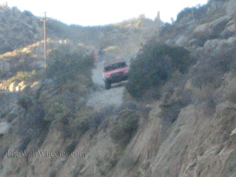 1990 Ford Bronco - red off-road race truck kicks up dust as it climbs a steep, rugged hillside trail among rocks and brush at dusk.