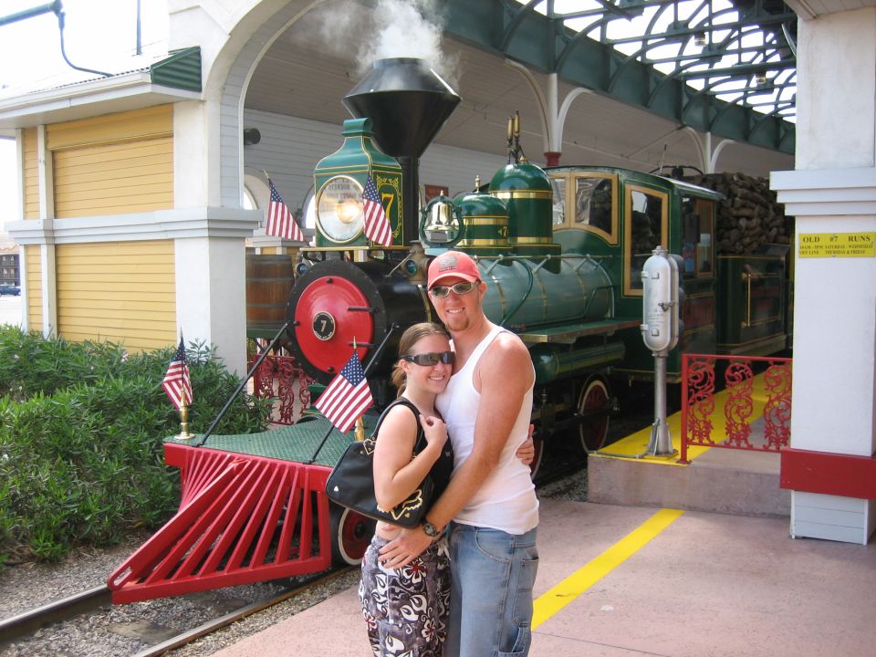Green locomotive numbered 7 with red cowcatcher and US flags at a covered station platform