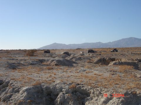 1996 Ford Bronco - Multiple off-road vehicles traverse a desert landscape, kicking up dust on a clear day.