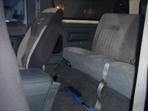 1989 Ford Bronco - Interior of a Ford truck looking rearward past a tilted-forward front bucket seat wearing a gray and black Ford-logo seat cover, toward the…