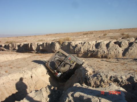 1996 Ford Bronco - An off-road vehicle is stuck in a deep ditch in a dry, arid landscape under a clear blue sky.