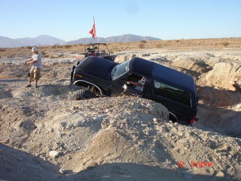 1996 Ford Bronco - A black SUV is stuck in a deep ditch off-road, with a person attempting to help and another vehicle in the background.