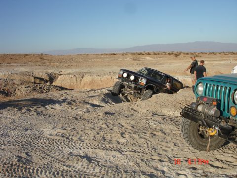 1996 Ford Bronco - A black Ford Bronco is stuck in a deep sandy ditch, with a teal Jeep attempting to pull it out in a desert environment.