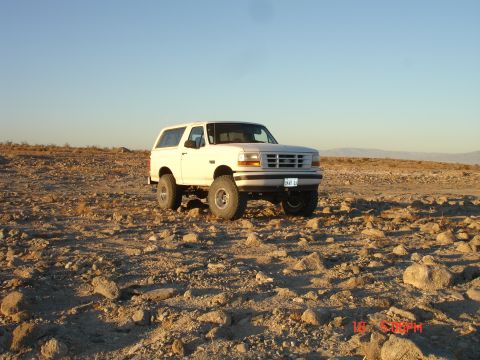 1996 Ford Bronco - Driver-side 3/4 front view of a white two-door Ford Bronco with hardtop parked on open rocky desert at golden hour, lifted on chrome-style…