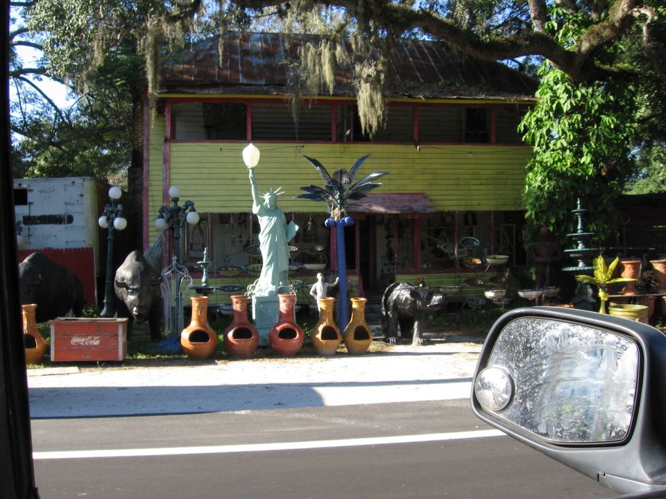 Yard sale with Statue of Liberty replica, animal statues, and pottery in front of a yellow house. Side mirror of a vehicle is in the foreground