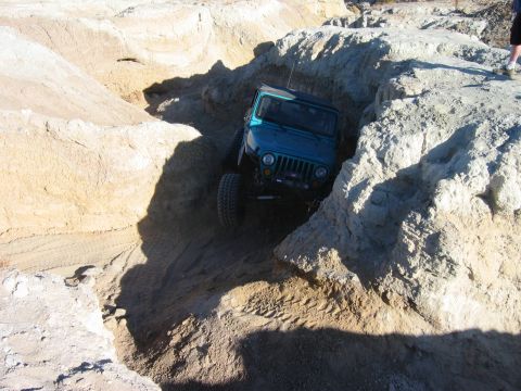 1985 Ford Bronco - A teal Jeep Wrangler maneuvers through a tight, sandy canyon with steep, eroded walls.