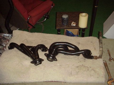 1989 Ford Bronco - A pair of black-coated tubular exhaust headers laid out on a white towel indoors, with a rusty EGR crossover tube and open-end wrenches bes…