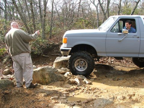 1996 Ford Bronco - A man directs a gray Ford Bronco as it drives over rocks and mud on an off-road trail.