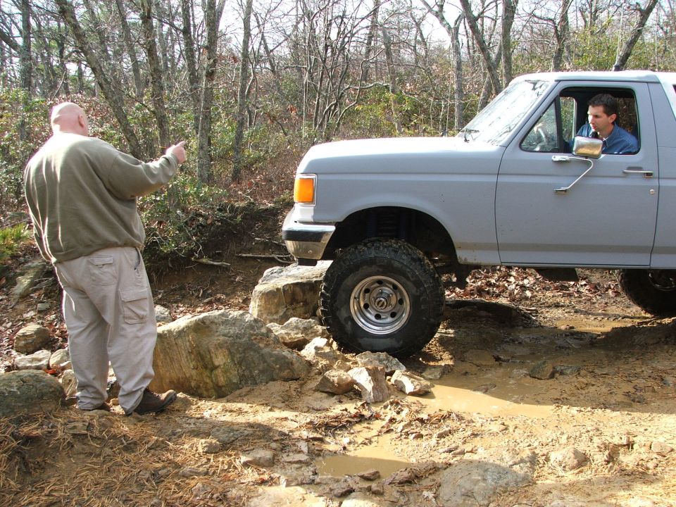 Gray 1992 Ford Bronco with large tires on a rocky, muddy trail, driver visible