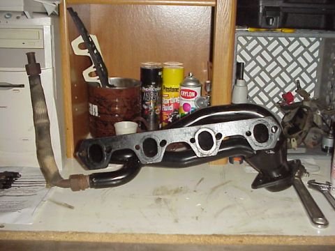1989 Ford Bronco - A black tubular exhaust header stood on end on a workbench, showing its four-port machined head flange and the merge collector below, with…