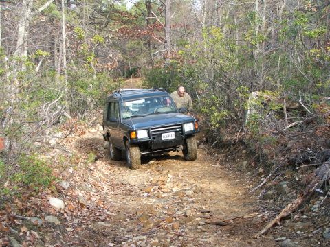 1996 Ford Bronco - A dark blue SUV navigates a challenging, rocky dirt path through a dense forest.