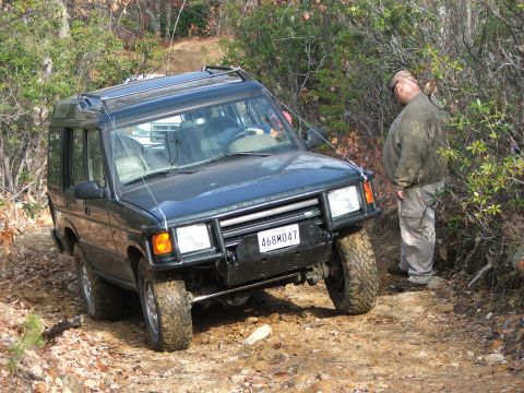 1996 Ford Bronco - Land Rover Discovery navigating a rough, wooded trail with a person standing nearby.