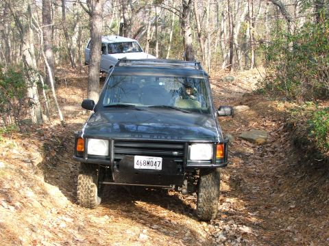 1996 Ford Bronco - Two SUVs navigate a wooded trail with fallen leaves covering the ground.