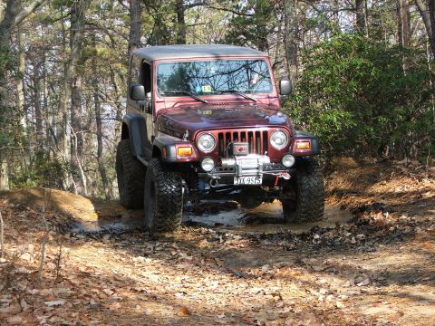 1996 Ford Bronco - A red Jeep Wrangler navigates a muddy path through a forest, its tires kicking up water and dirt.