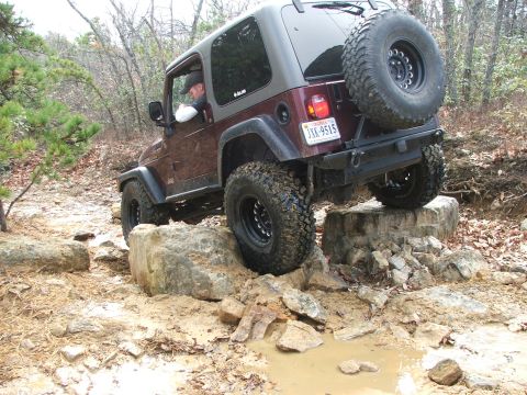 1996 Ford Bronco - A maroon Jeep Wrangler TJ tackles a challenging off-road course with large rocks and muddy puddles.