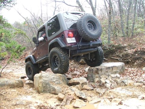 1996 Ford Bronco - A maroon Jeep Wrangler TJ navigates a rocky off-road course with its spare tire mounted on the back.