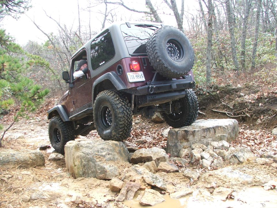 Maroon Jeep Wrangler TJ climbing over rocks on a trail, spare tire mounted on the rear