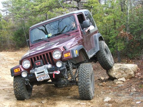 1996 Ford Bronco - A maroon Jeep Wrangler Rubicon navigates a rocky trail, with one wheel lifted high.