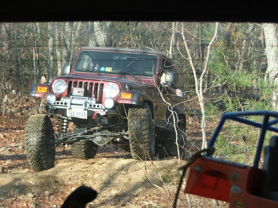 Red Jeep Wrangler with large tires on a dirt trail, another off-road vehicle in the foreground