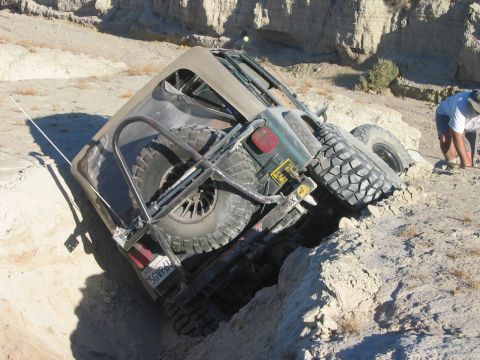 1985 Ford Bronco - A Jeep Wrangler is stuck tilted on its side in a rocky ditch, with a person nearby assessing the situation.