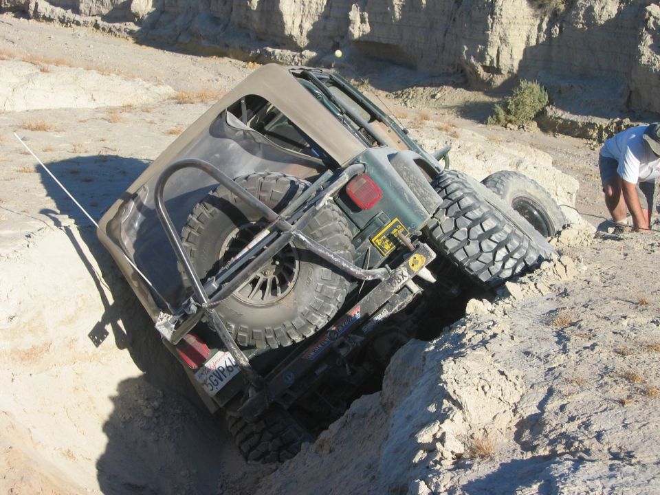 Green soft-top 4x4 tilted on rocks in desert with spare tire and mud tires, person at right