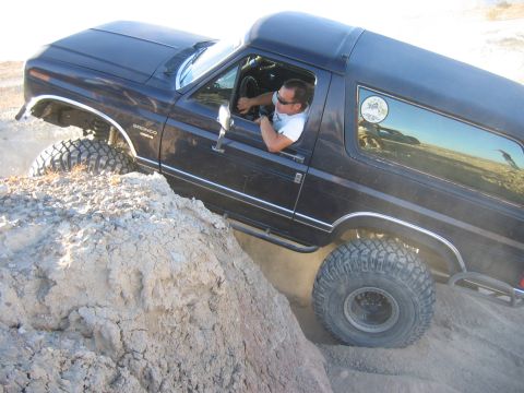 1985 Ford Bronco - A man drives a dark blue Ford Bronco up a steep dirt incline, showing off its off-road capabilities.