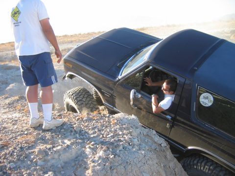 1985 Ford Bronco - A black 1985 Ford Bronco is shown off-roading on a rocky, uneven surface with a driver inside and a person standing nearby.