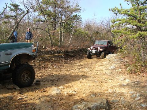 1996 Ford Bronco - Two off-road vehicles navigate a muddy and rocky path through a wooded area on a sunny day.