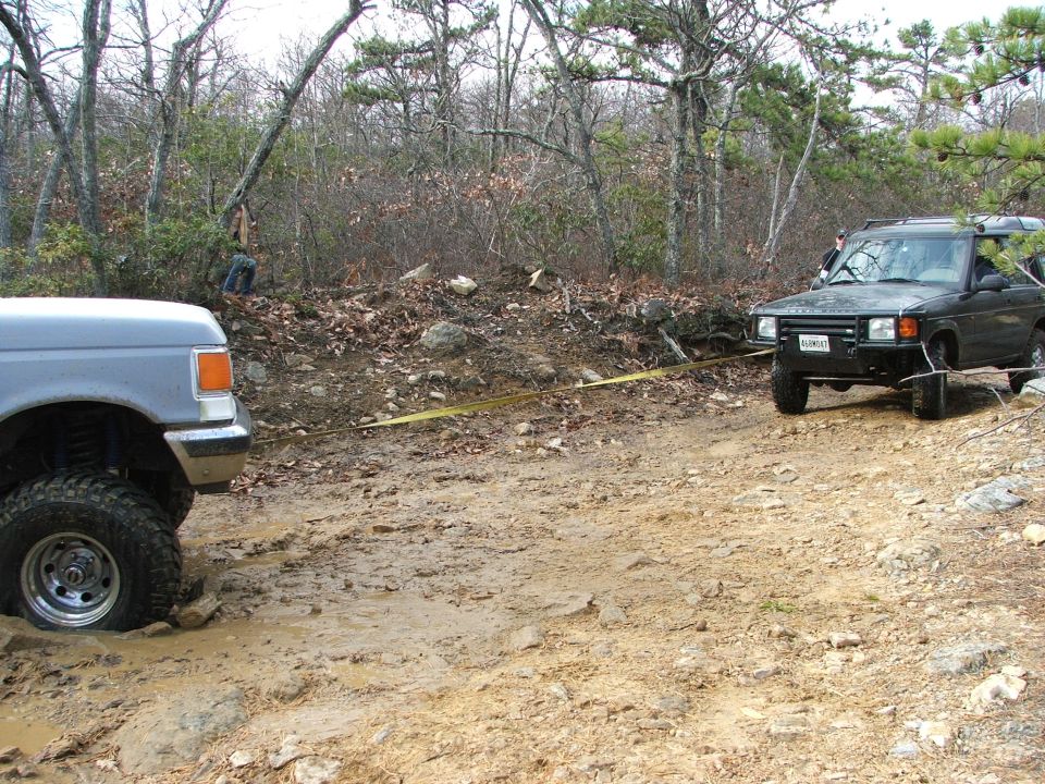 Gray 1996 Ford Bronco stuck in mud, being pulled by a dark green Land Rover Discovery on a dirt path