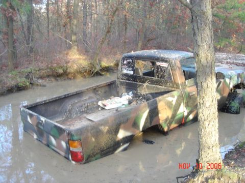 1992 Toyota Tacoma - Pickup truck with camouflage paint is stuck in muddy water in a wooded area.