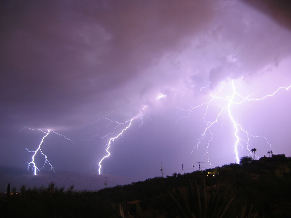 Lightning strikes during a thunderstorm over a dark, hilly landscape with some lights visible