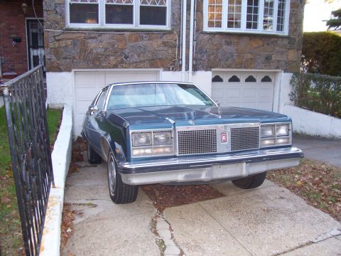 1999 Ford Crown Victoria - A blue 1977 Oldsmobile Delta 88 sedan is parked in front of a stone-clad house.