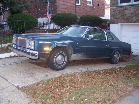 1999 Ford Crown Victoria - A dark blue 1977 Oldsmobile Cutlass Supreme is parked in a driveway.