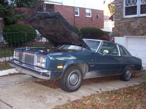 1999 Ford Crown Victoria - A vintage Oldsmobile coupe sits in a driveway with its hood up, revealing the engine bay.