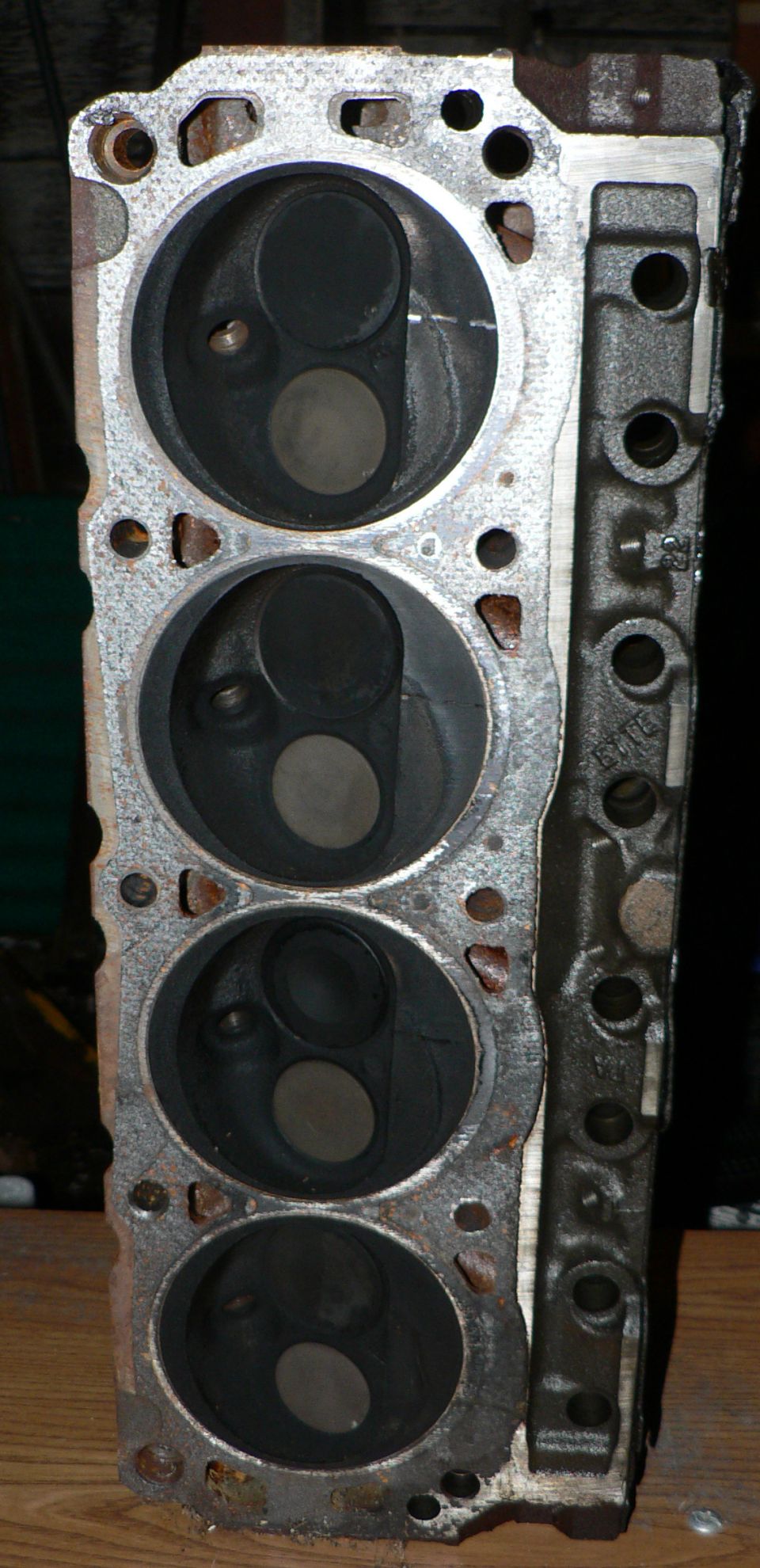 Bare cast-iron V8 cylinder head on wood bench, deck side up showing four combustion chambers