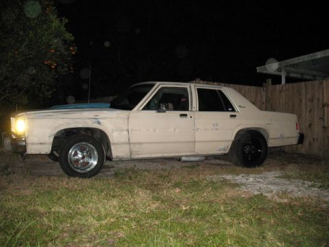 1985 Ford LTD Crown Victoria - A tan sedan sits on a grassy lawn at night, showing its side profile with modified wheels and exhaust pipes.